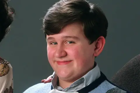 Uśmiechnięty Dudley Dursley, kuzyn Harry'ego Pottera, w niebieskim swetrze.